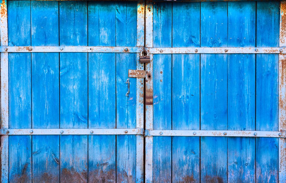 The old large wooden doors painted in blue and closed on the padlock. The blue gate on the lock. Wooden gates closeup, for background.